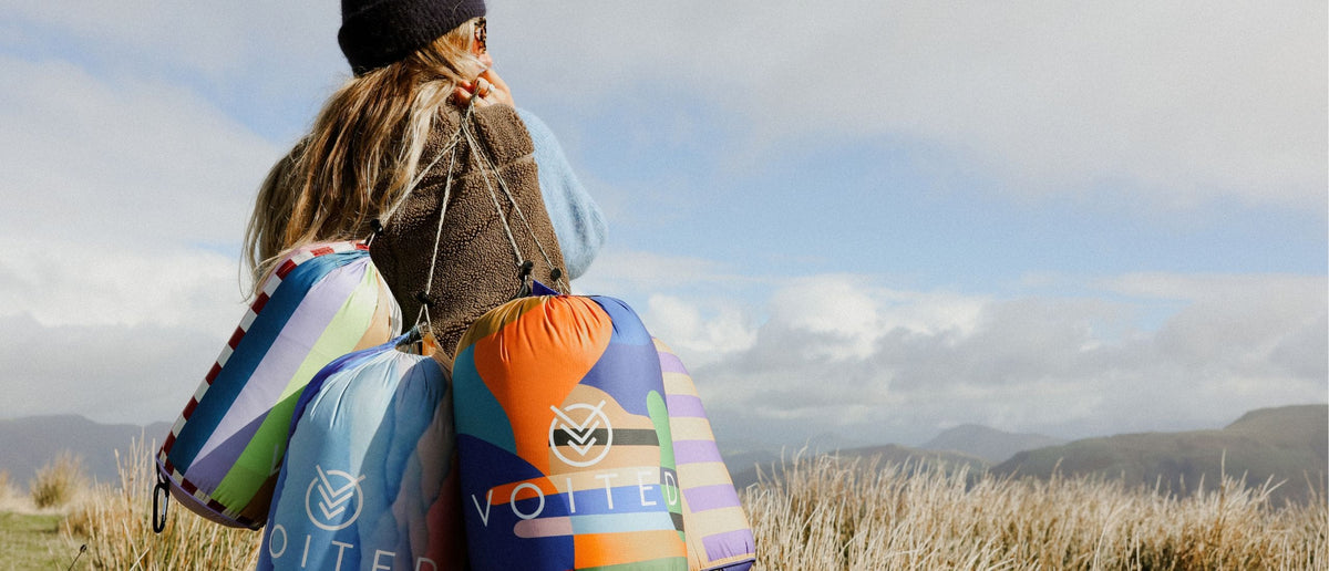 Woman carrying four colorful VOITED outdoor blankets in stuff sacks over her shoulder, with grassland and mountains behind.