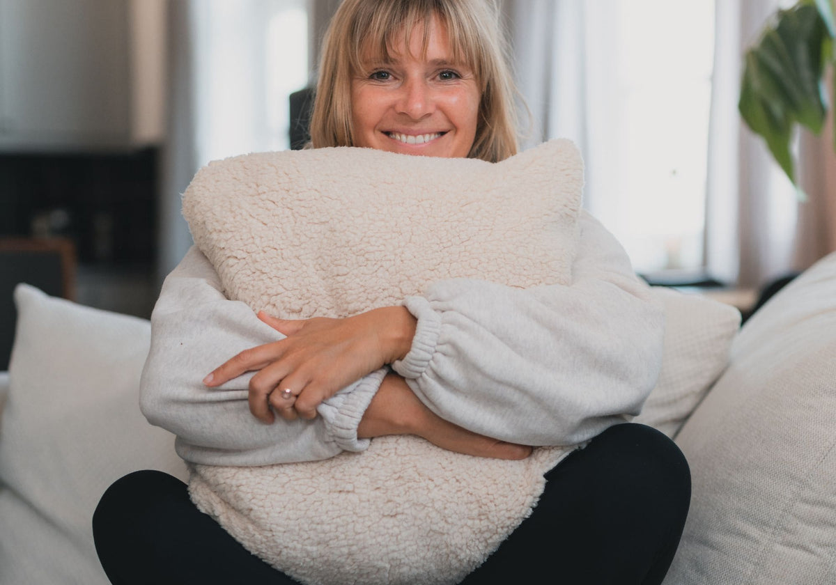 A smiling woman sits on a sofa hugging a large cream sherpa throw pillow. She is wearing a light gray knit sweater and black pants in a brightly lit living room.Cloudtouch VOITED