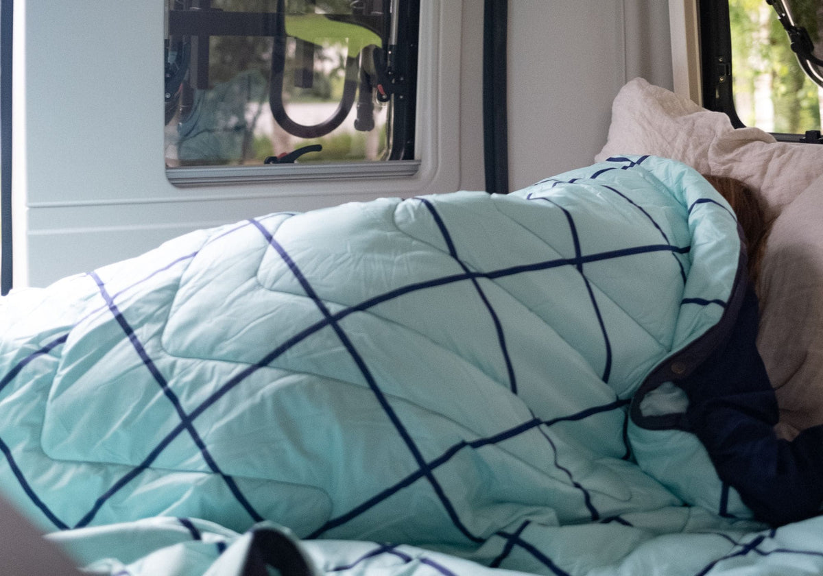 A light aqua quilted comforter with a dark navy grid pattern, shown on a bed with a person partially covered underneath, next to a beige pillow. VOITED POOL