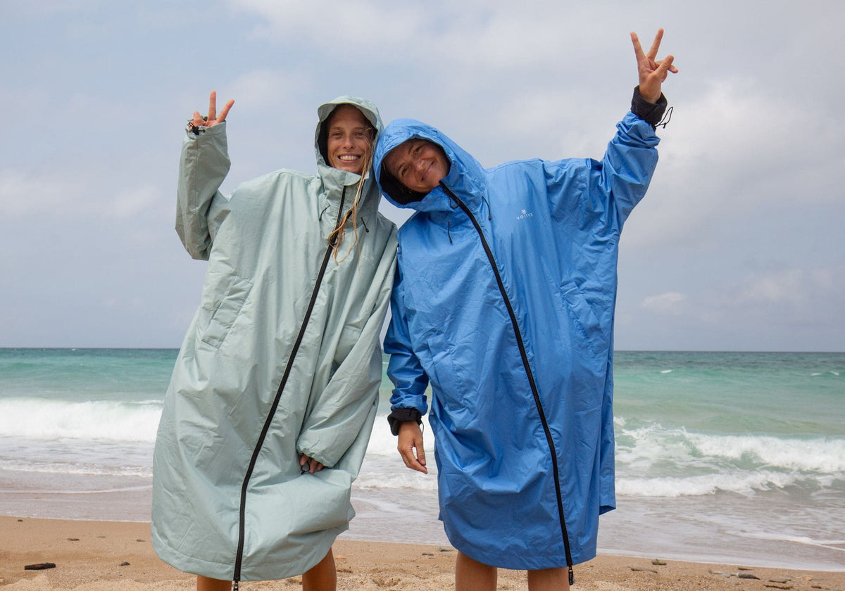 Two smiling women in VOITED Allweather Coats in sage green and blue, hoods up, making peace signs on a beach with waves behind them.