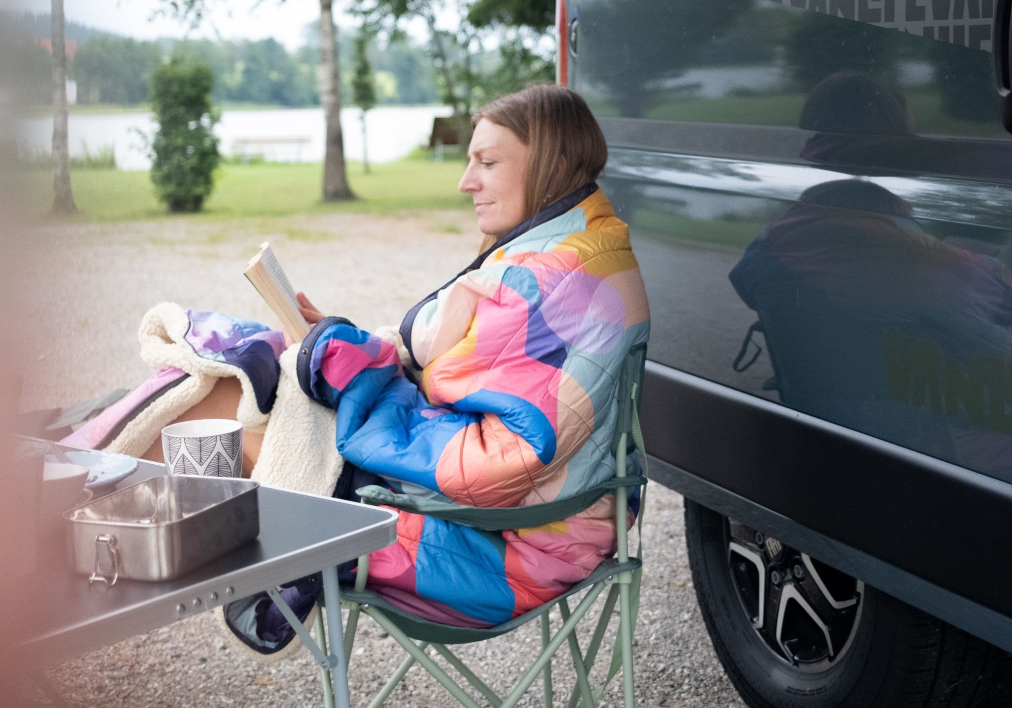 A woman sits in a green folding camp chair beside a black vehicle, wrapped in a colorful quilted blanket Cloudtouch VOITED and reading a book. A small table with a metal container is in the foreground, and trees and water are visible in the background.