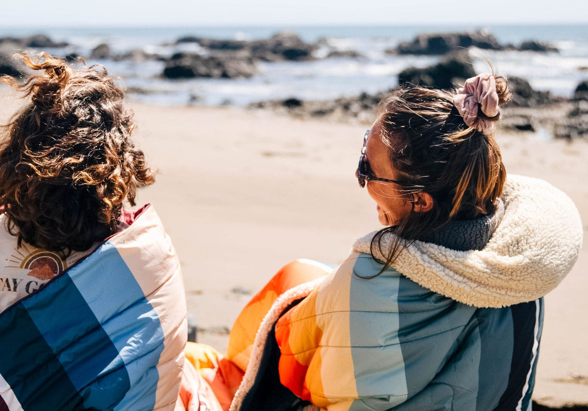 Two friends sit on a sunny, rocky beach wrapped in VOITED Cloudtouch blankets with colorful retro stripes in blue, teal, orange and cream, one with a cozy sherpa fleece hood, smiling as they look out at the sea.