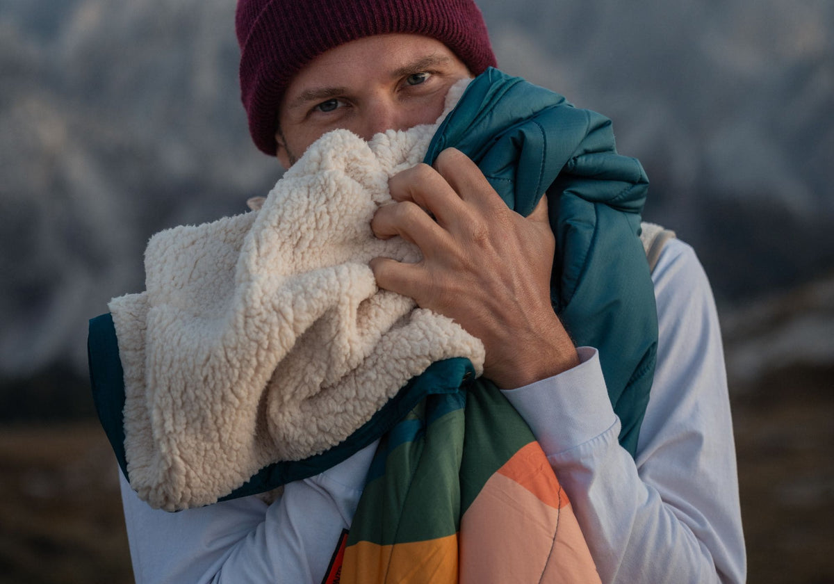 A man outdoors in a mountain setting, wearing a burgundy beanie, holds up a VOITED Cloudtouch Blanket with a cream sherpa fleece side and a teal and orange side, with mountains blurred in the background.
