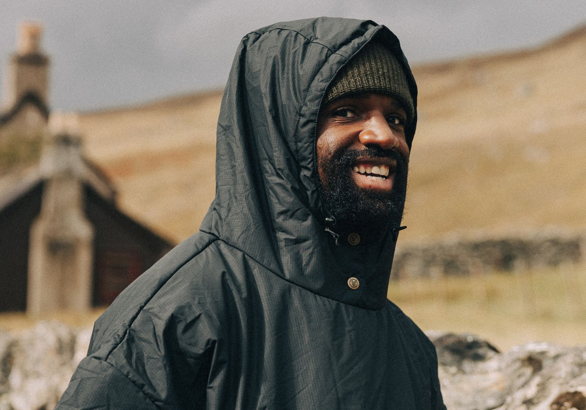 A smiling man wearing a dark green VOITED poncho with hood up over a beanie, standing outdoors in front of a rolling hillside and a stone cottage