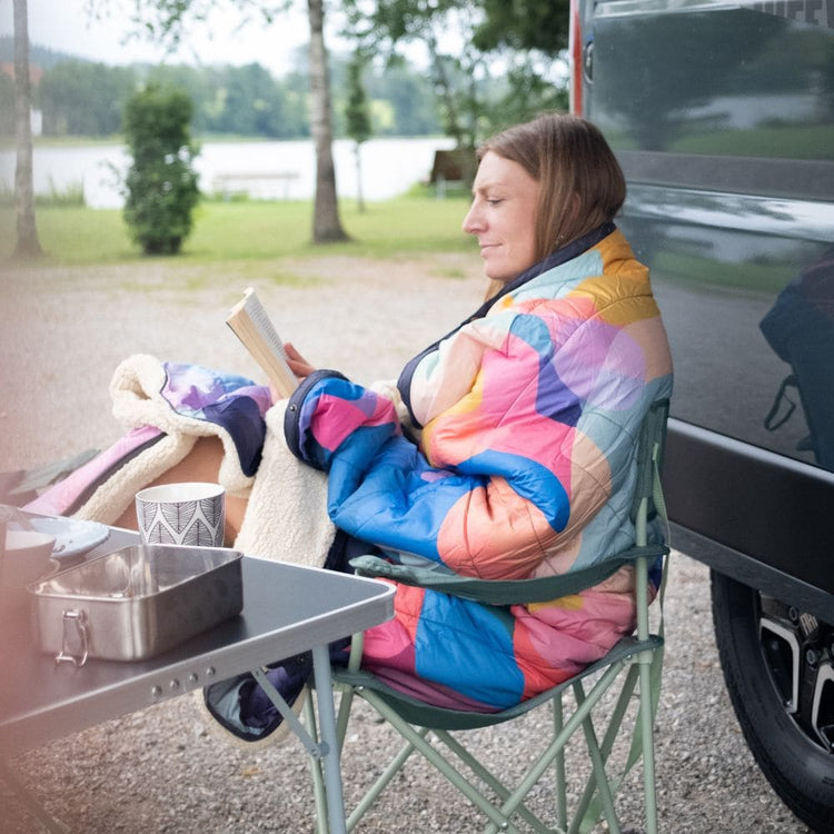 A woman sits in a green folding camp chair beside a black vehicle, wrapped in a colorful quilted blanket Cloudtouch VOITED and reading a book. A small table with a metal container is in the foreground, and trees and water are visible in the background.