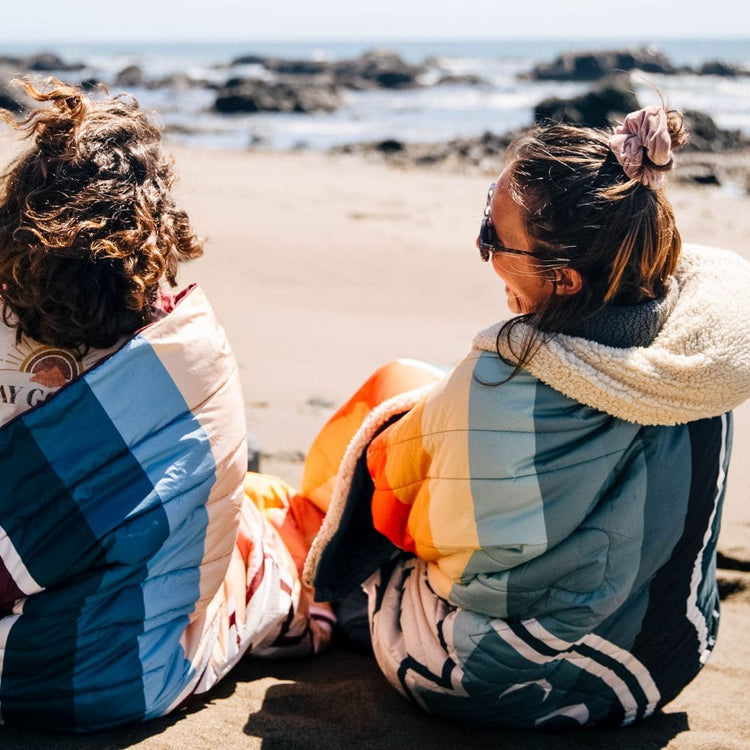 Two friends sit on a sunny, rocky beach wrapped in VOITED Cloudtouch blankets with colorful retro stripes in blue, teal, orange and cream, one with a cozy sherpa fleece hood, smiling as they look out at the sea.