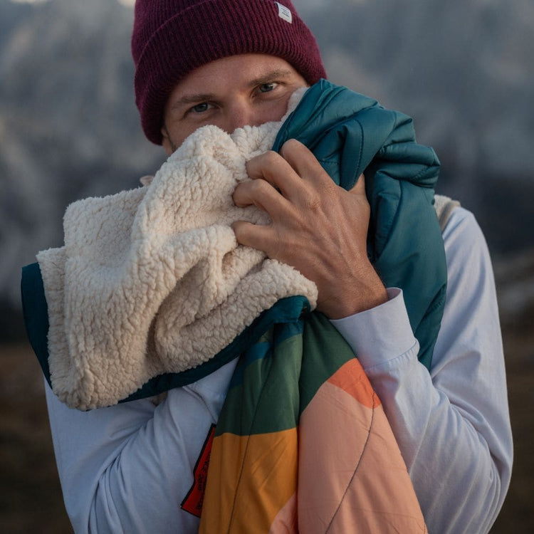 A man outdoors in a mountain setting, wearing a burgundy beanie, holds up a VOITED Cloudtouch Blanket with a cream sherpa fleece side and a teal and orange side, with mountains blurred in the background.