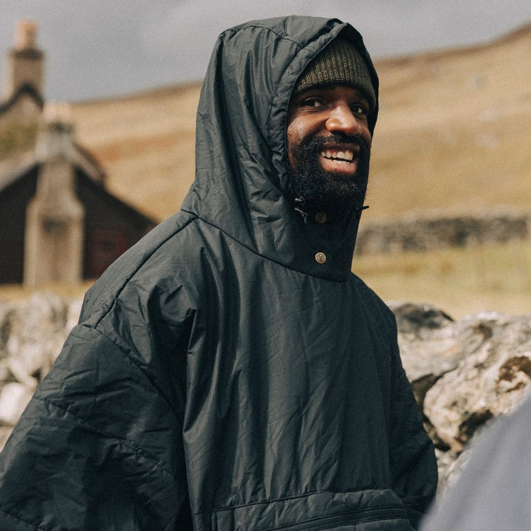 A smiling man wearing a dark green VOITED poncho with hood up over a beanie, standing outdoors in front of a rolling hillside and a stone cottage