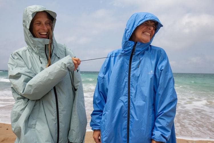 Two people wearing lightweight hooded rain jackets - All weather Coat stand on a beach, laughing while one pulls on the drawcord of the other jacket. The jacket on the left is a pale mint green and the jacket on the right is a medium blue.