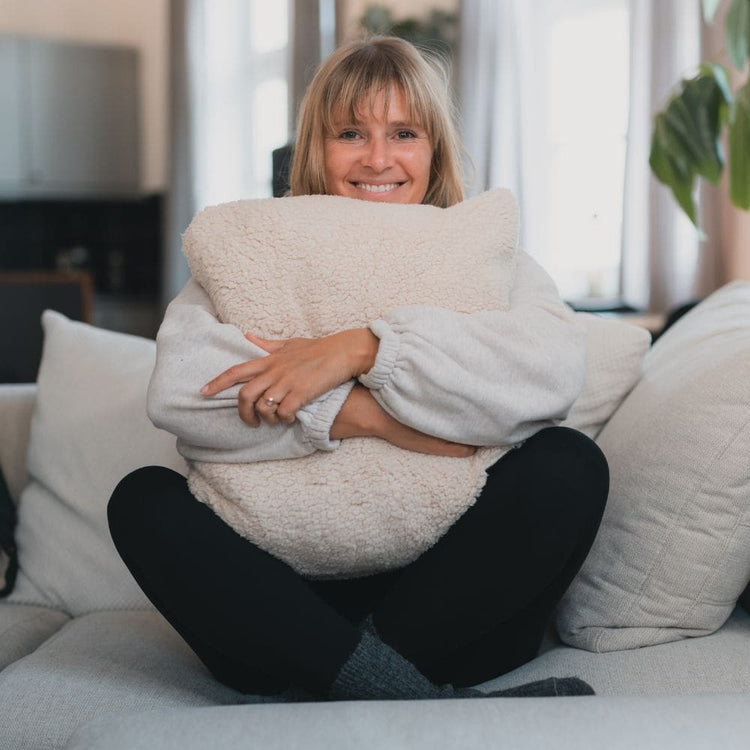 A smiling woman sits on a sofa hugging a large cream sherpa throw pillow. She is wearing a light gray knit sweater and black pants in a brightly lit living room.Cloudtouch VOITED