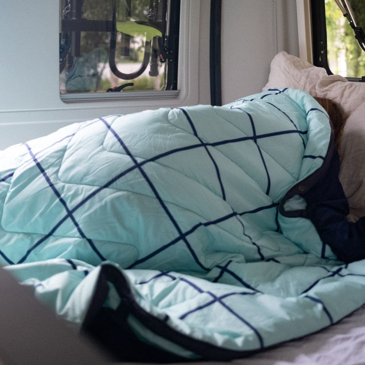 A light aqua quilted comforter with a dark navy grid pattern, shown on a bed with a person partially covered underneath, next to a beige pillow. VOITED POOL