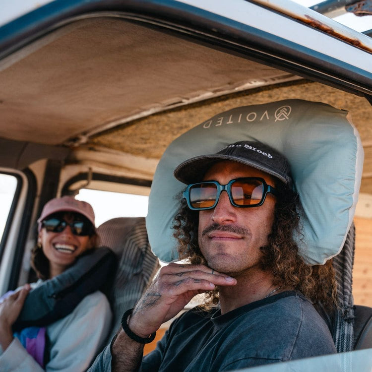 A man sitting in the driver's seat of a vehicle wearing a light blue Voited NeckPackTravel Blanket VOITED under his cap, with a woman smiling in the background. 