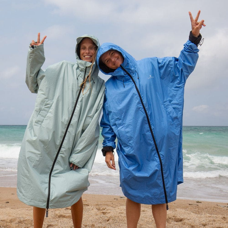 Two smiling women in VOITED Allweather Coats in sage green and blue, hoods up, making peace signs on a beach with waves behind them.